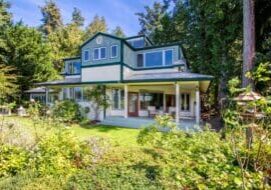 A two-story house with green trim, large windows, and a wraparound porch, surrounded by lush greenery and trees.