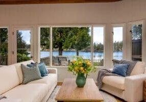 A living room with beige sofas, blue and green pillows, a wooden coffee table with a green vase of flowers, large windows offering a view of a lake and trees outside.