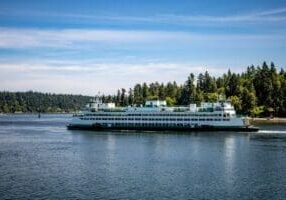 A large white and green ferry travels across a calm body of water with a forested shoreline and a clear blue sky in the background.