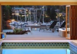 View from an indoor swimming pool overlooking a deck, with lounge chairs and a marina with boats docked on calm water, framed by sliding doors.