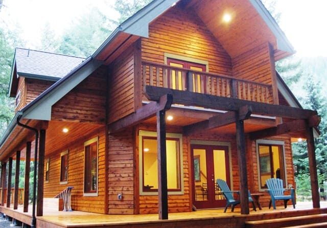 A two-story wooden cabin with a wraparound porch, pitched roof, and lit exterior lights sits amidst a forested background. Blue Adirondack chairs are placed on the porch.