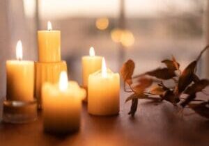 Several lit candles of varying sizes placed on a wooden surface with a blurred background and a sprig of leaves nearby.