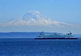 A ferry crosses a large body of water with a snow-capped mountain in the background under a clear blue sky.