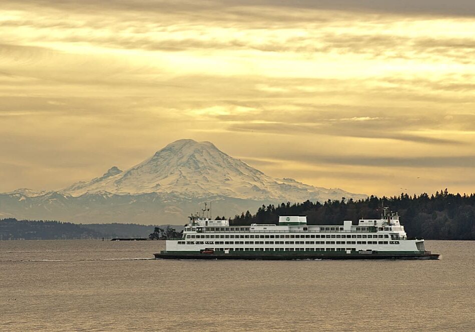 A ferry sails on calm waters against the backdrop of a towering snow-covered mountain under a cloudy sky.