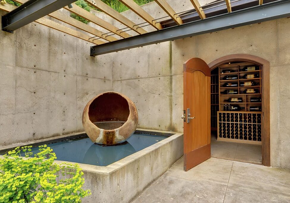 An open door reveals a wine cellar with numerous wine bottles on wooden shelves. Outside, there is a water feature with a large, hollow, spherical sculpture in a shallow pool.